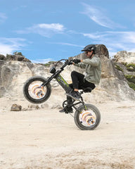 Person riding a small OTIDA R7 MAX Dual Motor Electric off-road bike on a rocky trail with a clear blue sky.