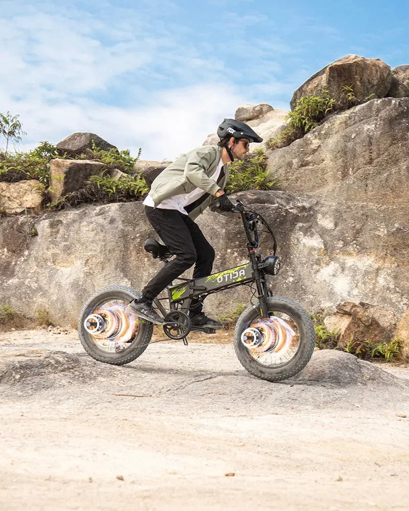 Person riding a small OTIDA R7 MAX Dual Motor Electric Bike on rocky terrain with a clear sky.
